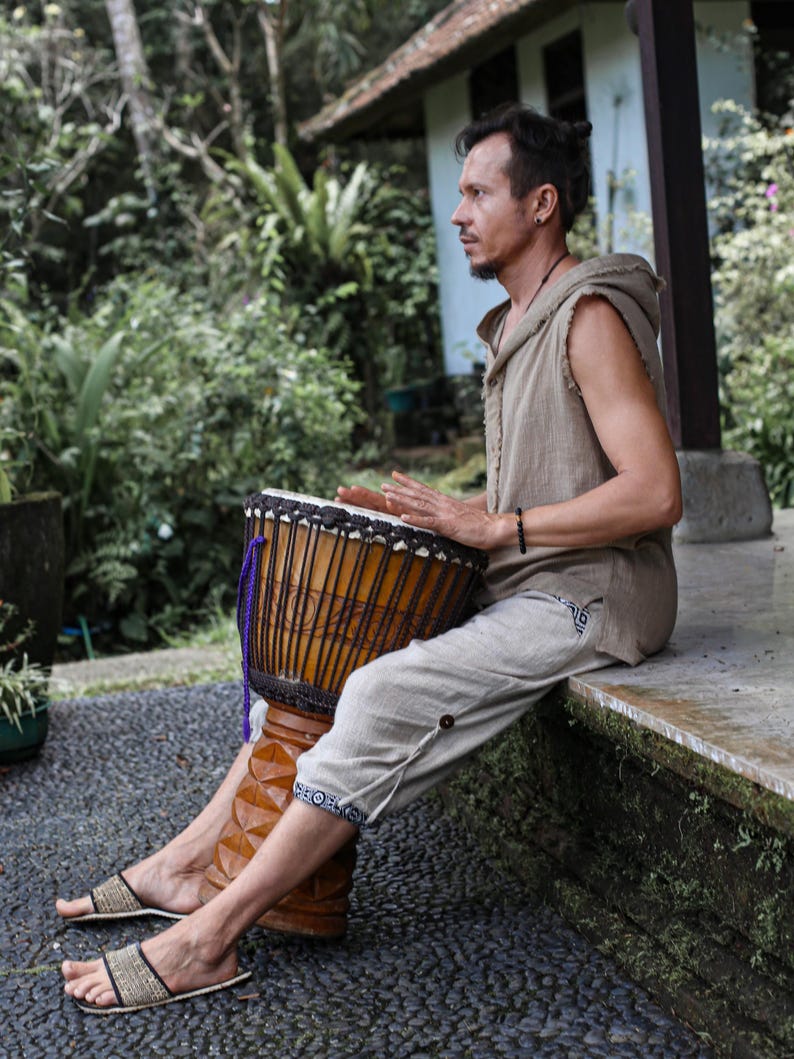 May include: A person playing a djembe drum outdoors. The drum is brown with vertical carved lines and a dark drumhead. The person is wearing a sleeveless tan shirt, light gray pants, and sandals. The background includes lush green foliage and a building.