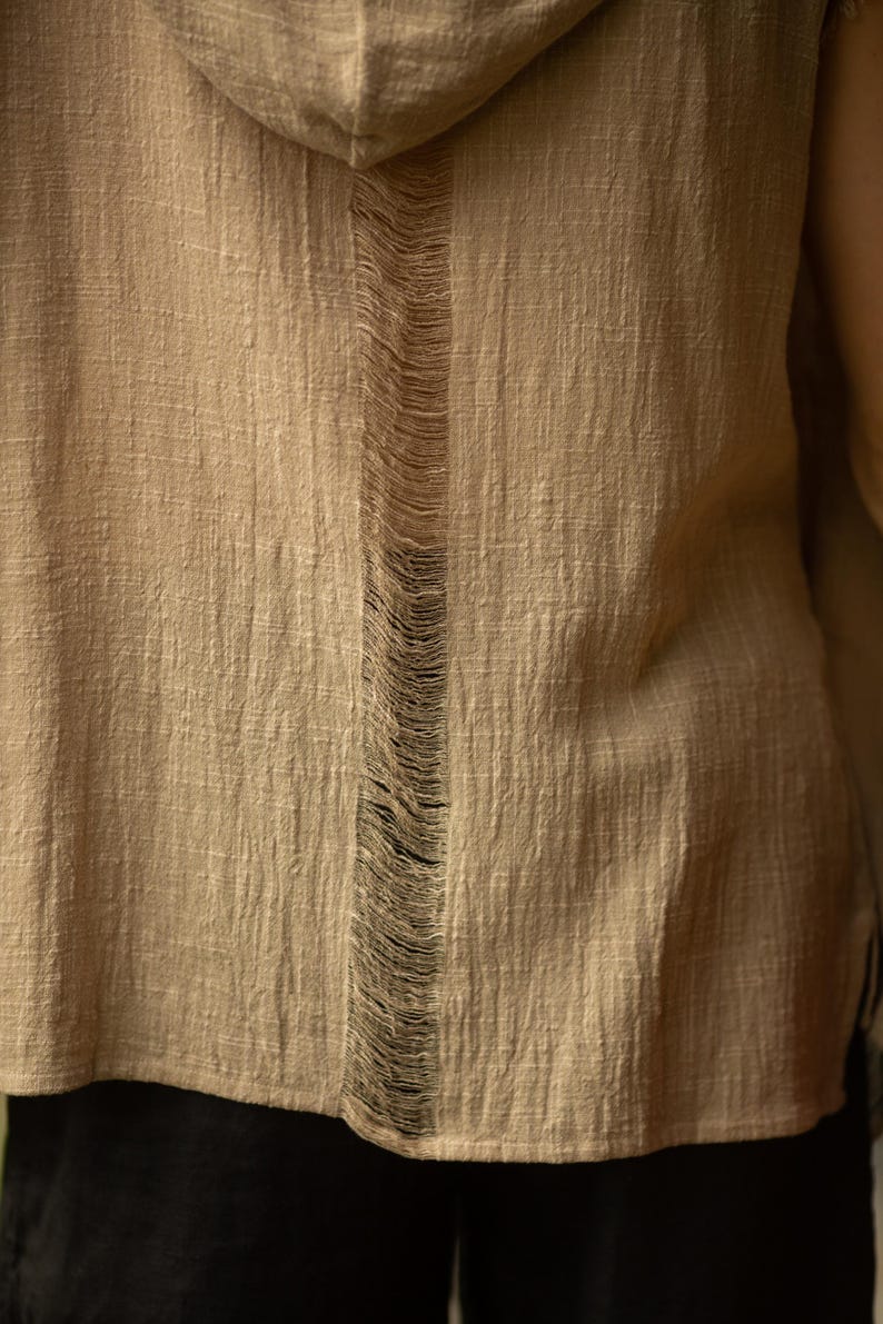 May include: A tan hooded shirt with a vertical distressed detail down the center back. The fabric has a textured appearance. The shirt's design is simple and casual, suitable for everyday wear. The hood is visible at the top of the image.
