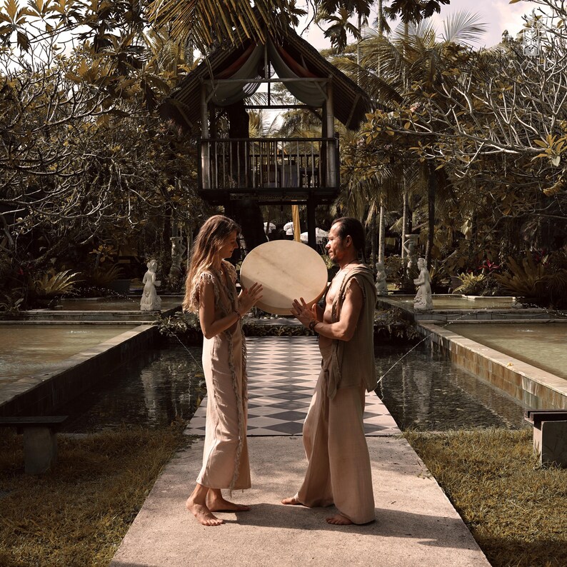 May include: Two people holding a large, round, light-colored drum outdoors. They are standing on a checkered path, with a gazebo in the background. The scene is set in a lush, green environment with a water feature.