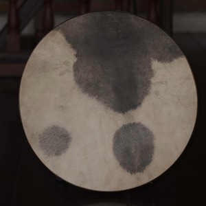 May include: A round, off-white drumhead with three dark gray patches. The drumhead is made of animal skin and has a natural, rustic look.