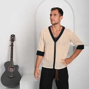 May include: A man wearing a white, semi-sheer shirt with black trim and a brown leather cord. He is wearing black pants and has a guitar in the background.