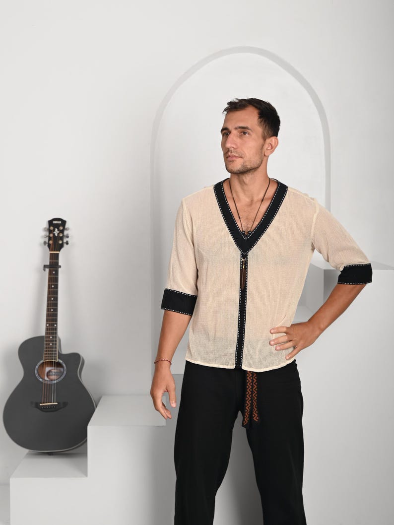 May include: A man wearing a white, semi-sheer shirt with black trim and a brown leather cord. He is wearing black pants and has a guitar in the background.