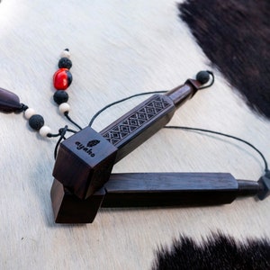 May include: Two dark brown wooden musical instruments, possibly percussion instruments, are displayed. One is on top of the other. The top instrument has a carved geometric design and the word "ayaho" with a leaf logo. A beaded strand with red, black, and white beads is attached.