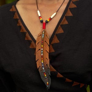 Op de afbeelding: Een leren ketting met een veerhanger in een verloop van bruin naar zwart. De veer is versierd met een centrale verticale lijn van veelkleurige kraaltjes. Het koord bevat witte, gele, rode en zwarte cilindrische kralen bij de bovenkant.