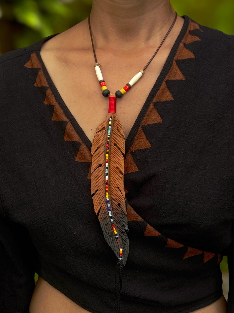 Op de afbeelding: Een leren ketting met een veerhanger in een verloop van bruin naar zwart. De veer is versierd met een centrale verticale lijn van veelkleurige kraaltjes. Het koord bevat witte, gele, rode en zwarte cilindrische kralen bij de bovenkant.