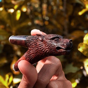 May include: A wooden pipe carved in the shape of an animal's head, with a brown and black finish. The pipe is held in a person's hand.