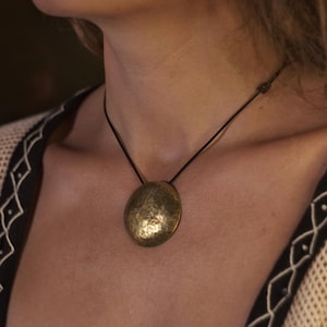 May include: A gold-toned hammered metal pendant necklace with a round disc shape, hanging from a black cord.