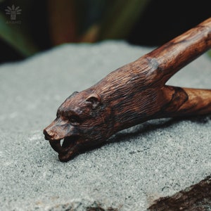 Può includere: Un fionda di legno scolpita a forma di testa d'orso. La fionda è di colore marrone scuro con venature del legno naturali. La testa dell'orso è dettagliata con una bocca spalancata e denti affilati.