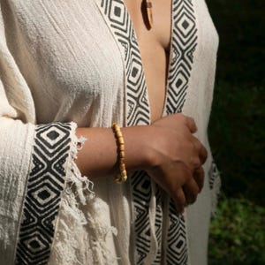 Könnte beinhalten: Cremefarbenes, offen geschnittenes Kleidungsstück mit schwarzem geometrischen Besatz. Ein Holzperlenarmband und eine Anhängerkette mit braunem Band sind sichtbar. Das Kleidungsstück hat einen Fransenrand und eine lockere Passform.