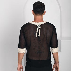 May include: A black sheer mesh shirt with three-quarter length sleeves and a cream-colored trim along the neckline and cuffs. The neckline features a decorative cream-colored tab with a geometric pattern and two hanging tassels at the center back.