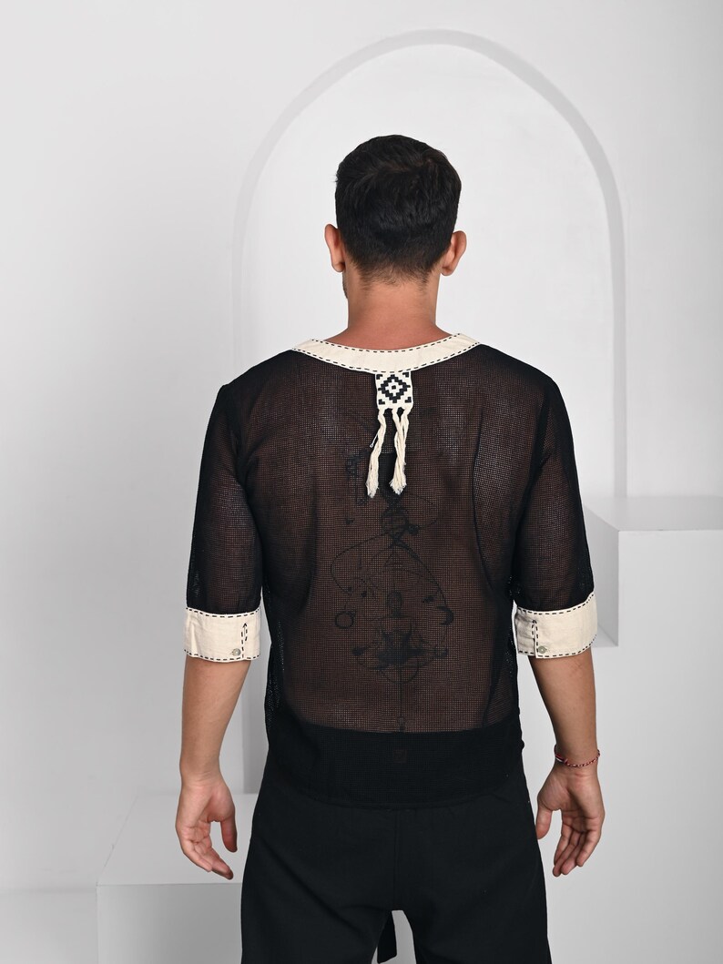 May include: A black sheer mesh shirt with three-quarter length sleeves and a cream-colored trim along the neckline and cuffs. The neckline features a decorative cream-colored tab with a geometric pattern and two hanging tassels at the center back.