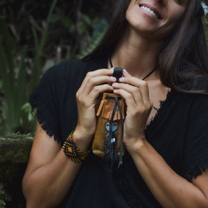 Peut inclure: Une femme tient une petite bourse en cuir marron avec des franges et un pompon perlé. La bourse a une sangle en cuir noir et est décorée d'un petit motif coloré. La femme porte une chemise noire et un bracelet perlé.