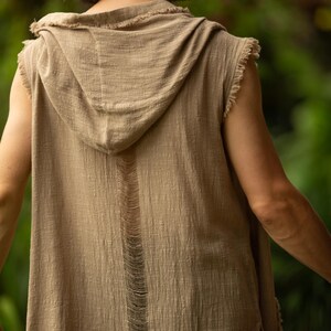 May include: A sleeveless, hooded top in a neutral tan color. The top features a distressed design with frayed edges and a vertical tear down the back. The fabric appears to be lightweight and textured, suitable for casual wear.