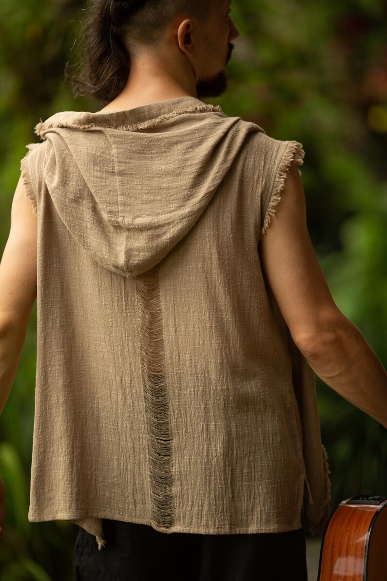 May include: A sleeveless, hooded top in a neutral tan color. The top features a distressed design with frayed edges and a vertical tear down the back. The fabric appears to be lightweight and textured, suitable for casual wear.