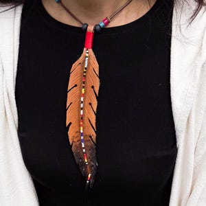 Op de afbeelding: Een leren veerhanger aan een ketting met een bruine en zwarte kleurverloop. De veer is versierd met een verticale lijn van veelkleurige kraaltjes in rood, geel, wit en blauw, hangend aan een bruin koord met kleine accentkralen.