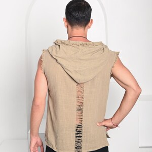 May include: A sleeveless, hooded shirt in a tan color with a distressed back design. The shirt has a raw edge finish on the hood and arm openings. The model is wearing black pants.