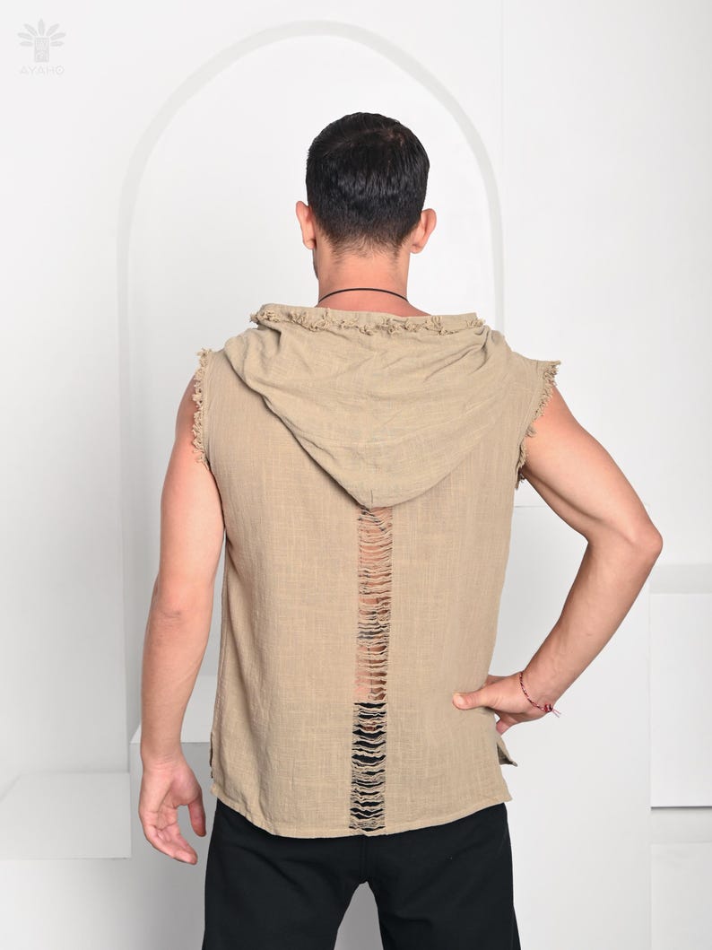 May include: A sleeveless, hooded shirt in a tan color with a distressed back design. The shirt has a raw edge finish on the hood and arm openings. The model is wearing black pants.
