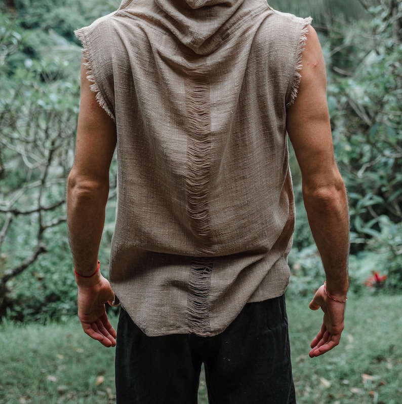 May include: A sleeveless, hooded top in a neutral tan color. The top features a distressed design down the back and frayed edges. The person is wearing black pants. The background is a lush green forest.