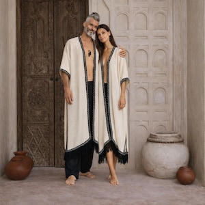 May include: Two matching cream-colored open-front robes featuring black geometric trim along the edges and black fringe detailing at the hem. The robes are long-sleeved and reach mid-calf length, worn over black trousers and a black skirt respectively.