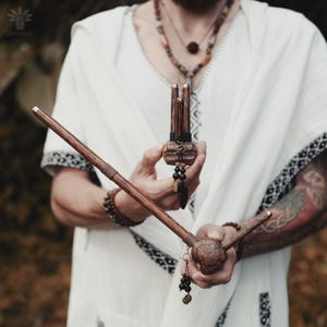 May include: Two dark brown wooden sticks with a polished finish and gold-toned metal accents. One stick is held horizontally, featuring a textured, rounded end with black beaded charms. The other is held vertically, featuring three parallel prongs.