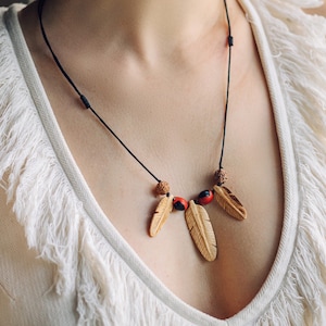 May include: A black cord necklace with three wooden feathers and red and black seed beads. The necklace is worn by a person with a white, fringed top.