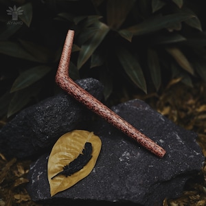 May include: A wooden smoking pipe with a brown and black speckled finish. The pipe is shaped like a boomerang and is resting on a rock.