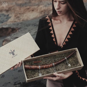 Könnte beinhalten: Eine zeremonielle Holzpfeife mit silbernen Akzenten wird in einer geflochtenen Schachtel mit einem braunen und grünen Logo präsentiert, auf dem "AYAHO" steht.