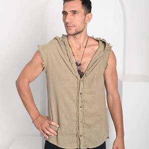 May include: A sleeveless, button-down hooded shirt in a light tan color. The shirt has a frayed edge design along the seams and a relaxed fit. The model is wearing black pants and a necklace. The background is white.