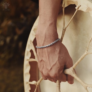 May include: A silver braided bangle bracelet on a person's wrist. The bracelet is simple and elegant, with a subtle braided design.