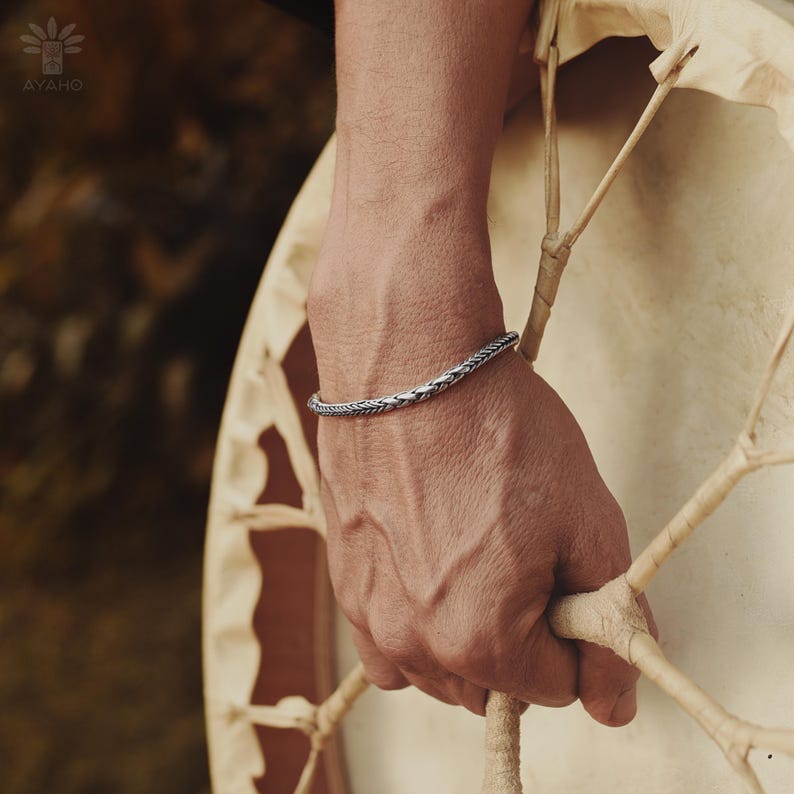 May include: A silver braided bangle bracelet on a person's wrist. The bracelet is simple and elegant, with a subtle braided design.