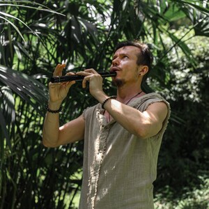 May include: A person playing a black flute outdoors, surrounded by lush green foliage. The person is wearing a sleeveless, button-down shirt. The flute is held with both hands, and the person is looking upwards.