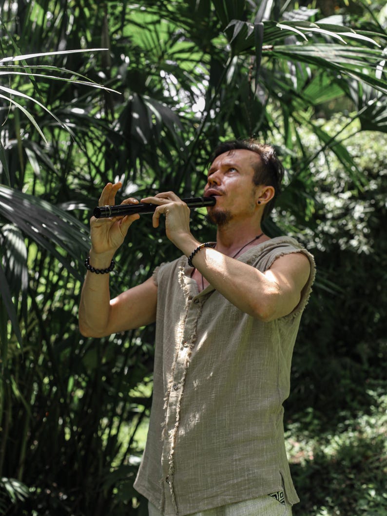 May include: A person playing a black flute outdoors, surrounded by lush green foliage. The person is wearing a sleeveless, button-down shirt. The flute is held with both hands, and the person is looking upwards.