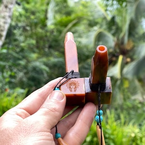 May include: A brown and tan pipe with a Native American design. The pipe has a rectangular base with a carved face and two cylindrical extensions. The pipe is decorated with black string and turquoise beads.