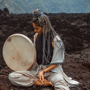 May include: A woman with long, braided gray hair sits on a dark, rocky surface. She holds a large, round, wooden frame drum with a light brown leather head. She is wearing a white, loose-fitting garment with black embroidery and a black top.
