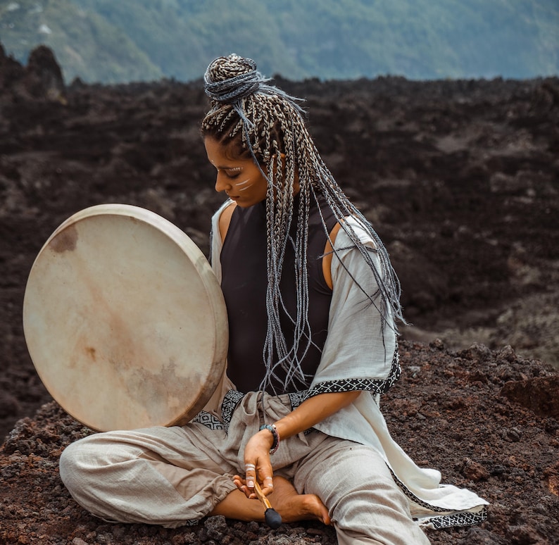 May include: A woman with long, braided gray hair sits on a dark, rocky surface. She holds a large, round, wooden frame drum with a light brown leather head. She is wearing a white, loose-fitting garment with black embroidery and a black top.