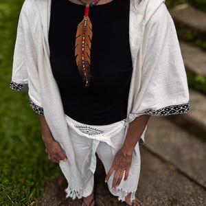 Op de afbeelding: Een zwart shirt met korte mouwen gecombineerd met een witte wikkelbroek met een zwarte oogafbeelding bij de taille. Een gebroken witte wikkel met capuchon en zwarte patroonrand wordt erover gedragen, aangevuld met een bruine leren verenketting.