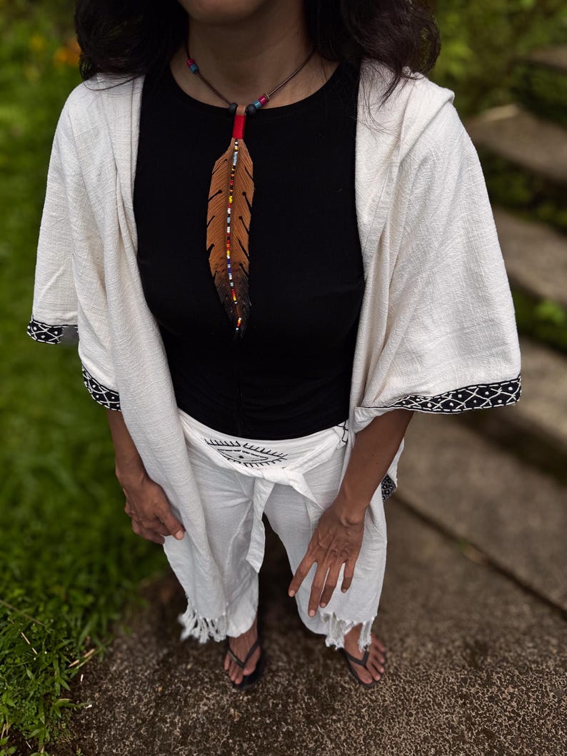 Op de afbeelding: Een zwart shirt met korte mouwen gecombineerd met een witte wikkelbroek met een zwarte oogafbeelding bij de taille. Een gebroken witte wikkel met capuchon en zwarte patroonrand wordt erover gedragen, aangevuld met een bruine leren verenketting.