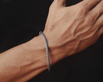 May include: A silver-toned bracelet featuring a dense, woven foxtail chain design. The metal has a polished finish with darkened recessed areas that highlight the intricate, repeating V-shaped pattern of the links throughout the entire band.