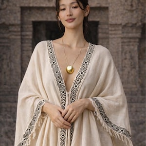 May include: A cream-colored, open-front caftan with black and white geometric trim. The caftan has long sleeves and a fringed hem. A gold pendant necklace with a large, round charm is worn with the caftan. The background is a stone wall.
