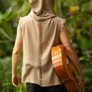May include: A person wearing a tan hooded sleeveless shirt with a distressed back design, holding a brown acoustic guitar. The guitar has a glossy finish. The person is wearing black pants. The background is a lush green garden.