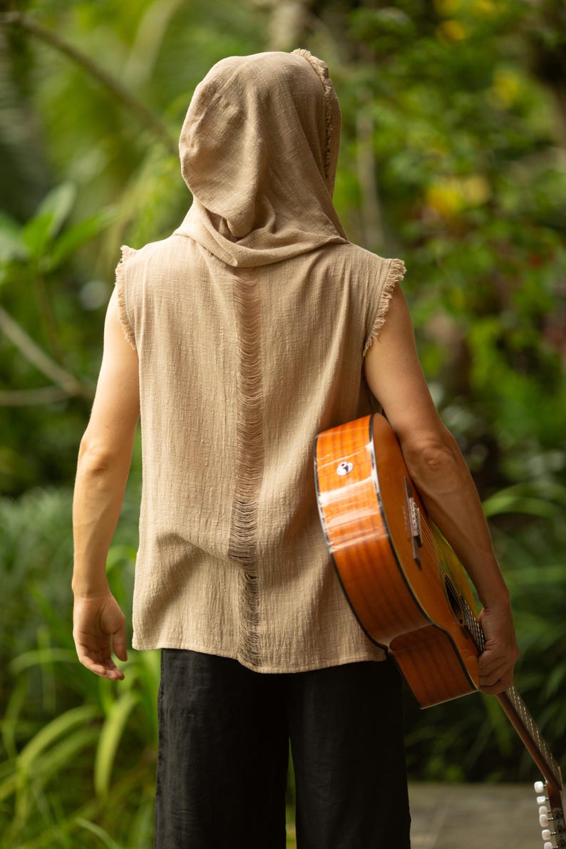 May include: A person wearing a tan hooded sleeveless shirt with a distressed back design, holding a brown acoustic guitar. The guitar has a glossy finish. The person is wearing black pants. The background is a lush green garden.
