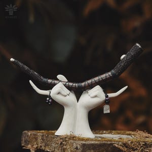Puede incluir: Una pipa de madera curvada con intrincados grabados negros en ambos extremos y un centro marrón oscuro envuelto en cordón negro. La pipa descansa sobre una escultura blanca de dos manos unidas, con pequeños dijes colgantes de cuentas oscuras.