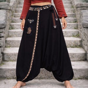 May include: Black harem pants featuring a drop-crotch design, a wide waistband with a geometric patterned trim, and a side pocket adorned with a hand symbol graphic. The pants include a matching patterned fabric tie and decorative metal accents.