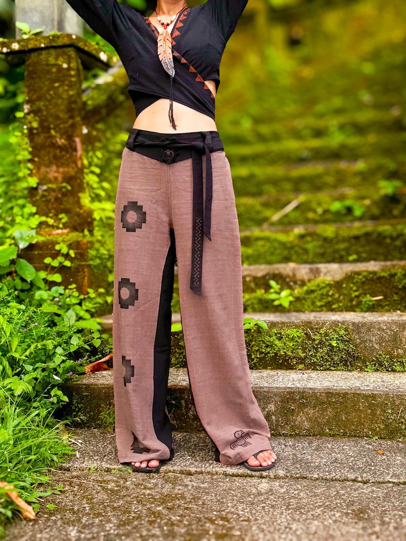 Op de afbeelding: Een zwarte overslagtop met V-hals en bruine geometrische rand, gecombineerd met een wijde broek. De broek is bruin met een zwarte tailleband, een zwarte strikceintuur en zwarte geometrische en vogelvormige prints op de onderkant.