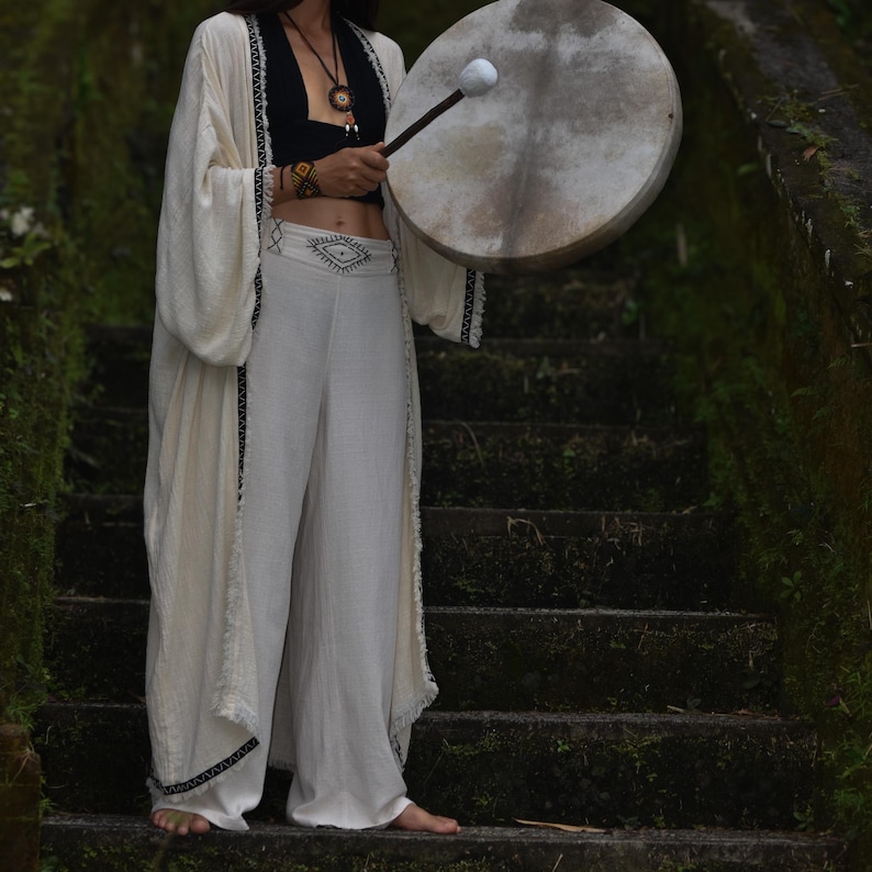 May include: A person wearing a white, crocheted, long-sleeved robe with black trim and white pants. They are holding a large, round, wooden drum with a white mallet.