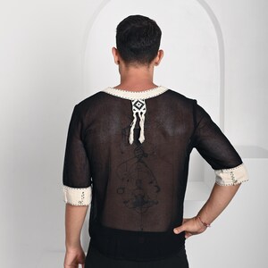 May include: A black mesh shirt with half-length sleeves, featuring cream-colored trim on the neckline and cuffs. The neckline includes a decorative cream and black woven tab with dangling fringe. The fabric is semi-sheer with a visible grid texture.