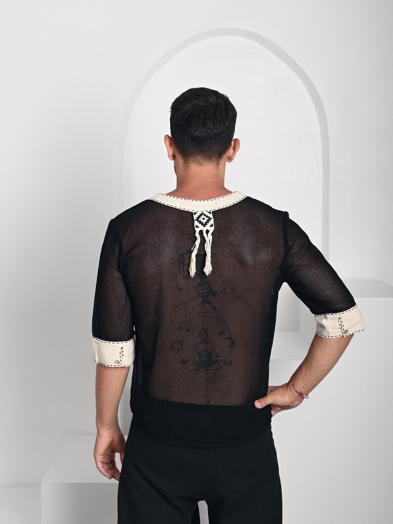 May include: A black mesh shirt with half-length sleeves, featuring cream-colored trim on the neckline and cuffs. The neckline includes a decorative cream and black woven tab with dangling fringe. The fabric is semi-sheer with a visible grid texture.