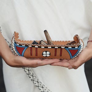 Peut inclure: Un plateau en forme de canoë en cuir peint à la main, avec des accents rouges, bleus et blancs. Le plateau contient des objets en bois et est tenu par une personne vêtue d'un vêtement blanc. Le plateau a un design décoratif.