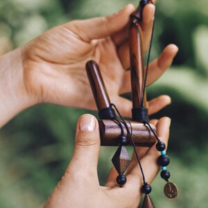 May include: A handcrafted wooden incense holder with a dark finish. The holder features a geometric design with black string and beads, including a diamond-shaped pendant and a small coin charm. The background is a soft green.