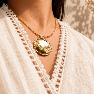 May include: A gold-toned necklace with an oval pendant. The pendant has a reflective surface and is framed by a twisted rope design. The necklace chain is also a twisted rope style. The background is a neutral color.